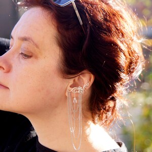 May include: Silver ear cuff with cascading chains and decorative filigree. The ear cuff features a floral design with dangling chains of varying lengths. The jewelry is worn on the ear and is a unique accessory.
