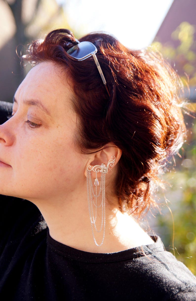 May include: Silver ear cuff with cascading chains and decorative filigree. The ear cuff features a floral design with dangling chains of varying lengths. The jewelry is worn on the ear and is a unique accessory.
