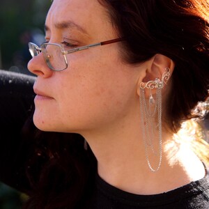May include: A silver ear cuff with a delicate, swirling design and dangling chains. The ear cuff is worn on the ear and features small, clear gemstones. The person in the image is wearing glasses and a black shirt.