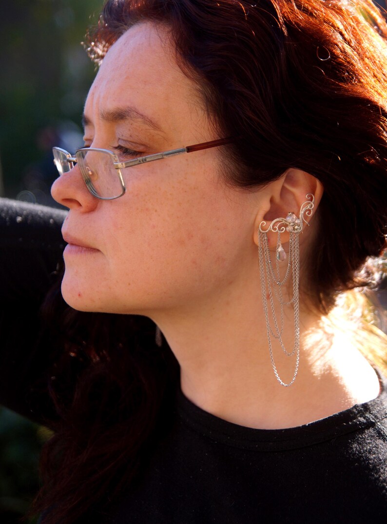 May include: A silver ear cuff with a delicate, swirling design and dangling chains. The ear cuff is worn on the ear and features small, clear gemstones. The person in the image is wearing glasses and a black shirt.