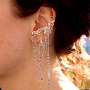 May include: Silver ear cuff featuring a delicate floral design with dangling chains. The ear cuff is adorned with small, clear gemstones and a pear-shaped crystal pendant. The jewelry is designed to be worn on the ear.