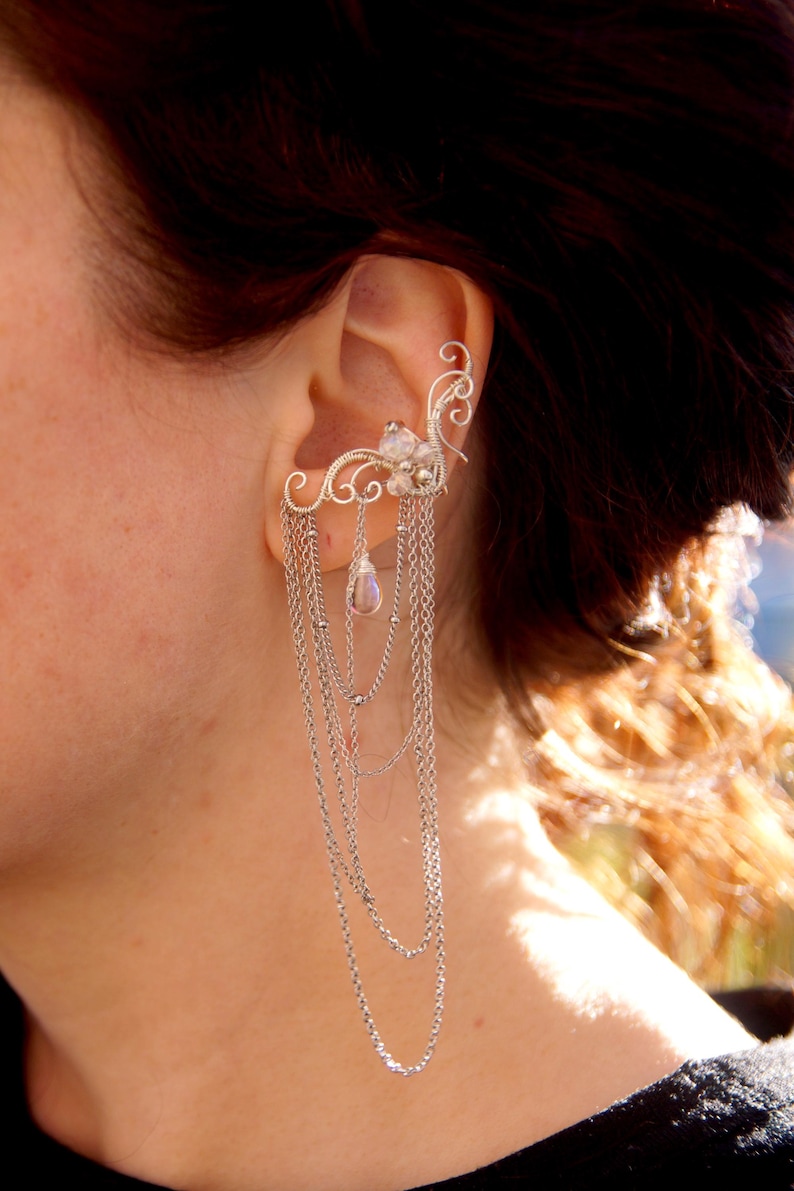 May include: Silver ear cuff featuring a delicate floral design with dangling chains. The ear cuff is adorned with small, clear gemstones and a pear-shaped crystal pendant. The jewelry is designed to be worn on the ear.