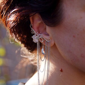 May include: A silver ear cuff with a floral design and dangling chains. The ear cuff features a cluster of clear crystals and a pear-shaped iridescent bead. The chains cascade down, creating a layered effect.