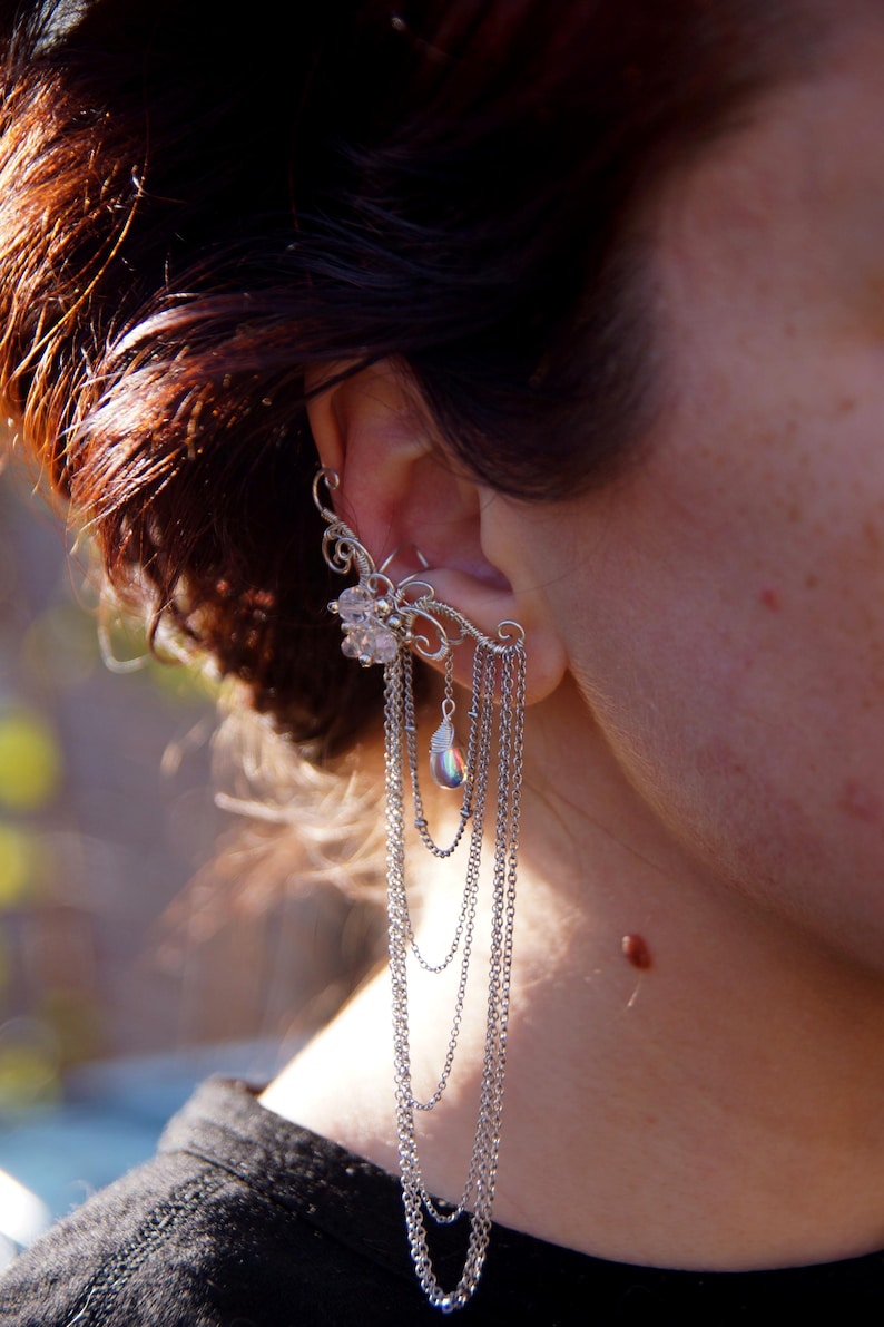 May include: A silver ear cuff with a floral design and dangling chains. The ear cuff features a cluster of clear crystals and a pear-shaped iridescent bead. The chains cascade down, creating a layered effect.