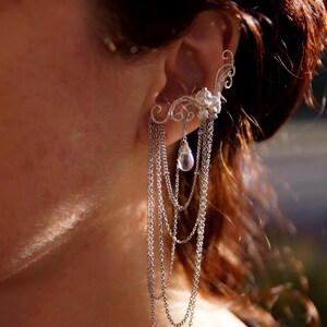 May include: A silver ear cuff with cascading chains and a floral design. The ear cuff features a cluster of clear crystals and a teardrop-shaped pendant. The jewelry is worn on the ear and dangles down.