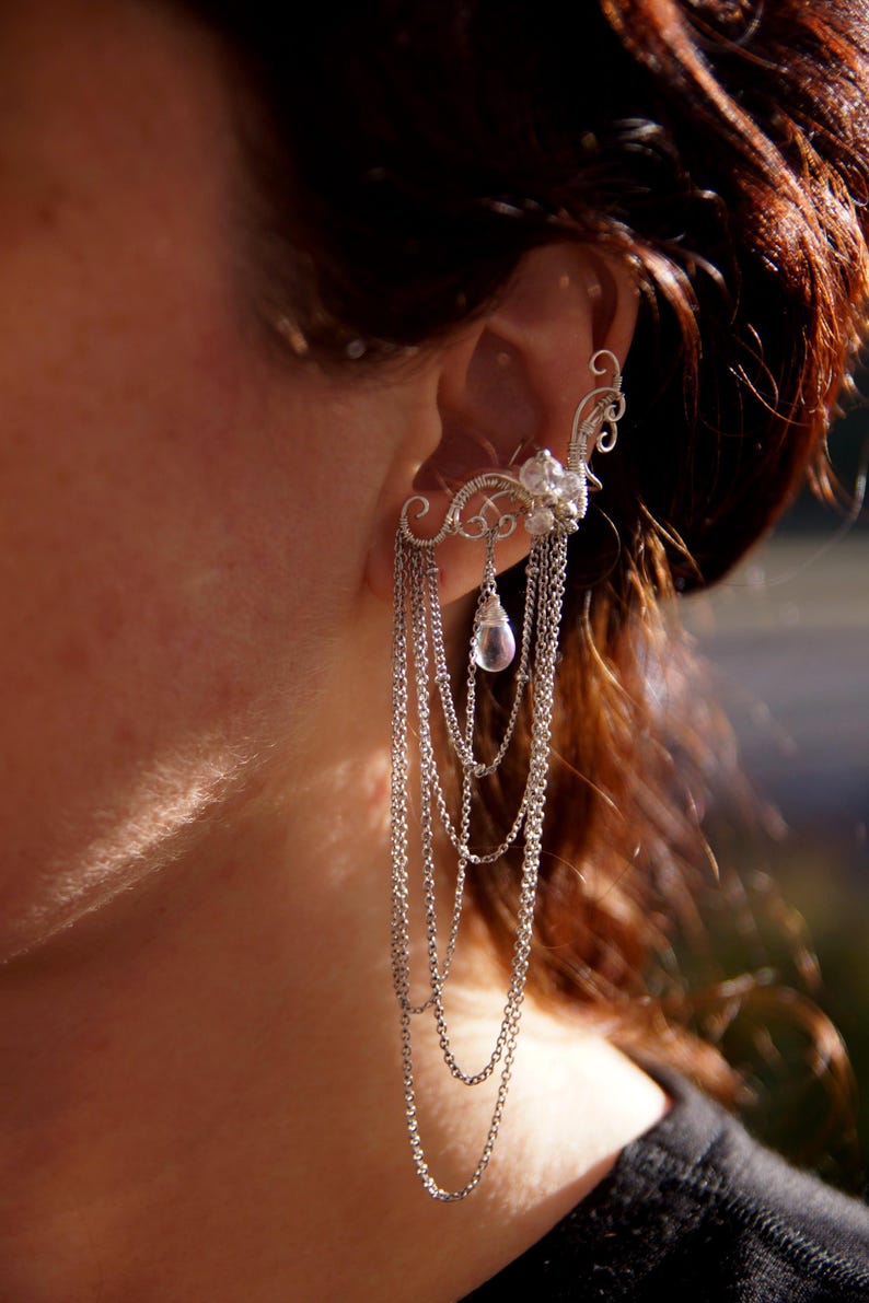 May include: A silver ear cuff with cascading chains and a floral design. The ear cuff features a cluster of clear crystals and a teardrop-shaped pendant. The jewelry is worn on the ear and dangles down.