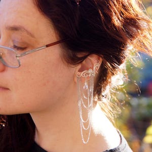 May include: A silver ear cuff featuring an ornate, swirling design that curves around the ear. Delicate chains cascade down, accented with a small, pearlescent bead. The jewelry is worn on the ear.
