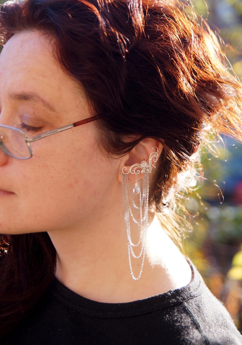 May include: A silver ear cuff featuring an ornate, swirling design that curves around the ear. Delicate chains cascade down, accented with a small, pearlescent bead. The jewelry is worn on the ear.