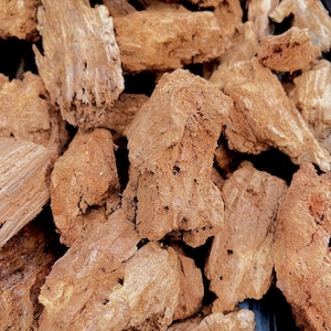 May include: A collection of rough, irregular pieces of light brown, porous wood bark. The fragments display a fibrous, weathered texture with deep crevices, jagged edges, and a dry, matte appearance, showing natural variations in wood grain.