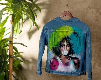 Puede incluir: Una chaqueta vaquera azul con una pintura colorida de una mujer con el pelo verde y azul soplando una burbuja en la espalda.
