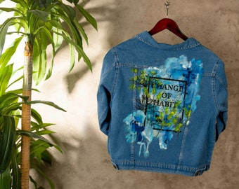 Könnte beinhalten: Eine blaue Jeansjacke mit einem bemalten Design auf der Rückseite. Das Design zeigt eine grüne Pflanze, blaue und weiße Aquarellspritzer und den Text "Change of Habit" in einem schwarzen Rahmen.
