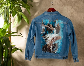 May include: A light blue denim jacket with a painted image of a ballerina in a white tutu. The artwork features a dancer in mid-leap with arms outstretched, set against a backdrop of blue and brown brushstrokes. The jacket is hanging on a wooden hanger.