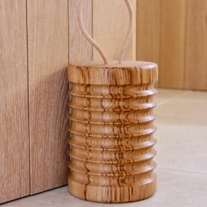 May include: A cylindrical wooden doorstop featuring a ribbed, textured surface and a light-colored wood grain finish. The top of the doorstop includes a looped handle made of a thin, flexible material for easy carrying and placement.