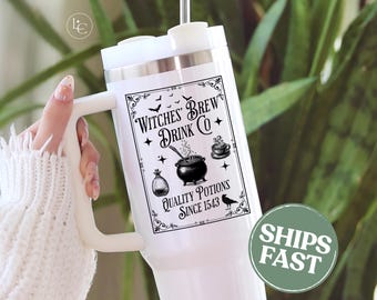 May include: White insulated tumbler with a handle, featuring a black and white graphic that reads "Witches Brew Drink Co." with illustrations of a cauldron, potion bottle, and raven. The tumbler has a silver rim and a white lid.