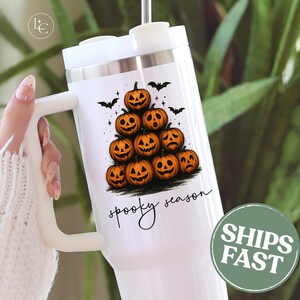 May include: White tumbler featuring a stack of orange jack-o'-lanterns and black bats, with the words "spooky season" in a handwritten font. The tumbler has a white handle and a silver rim.