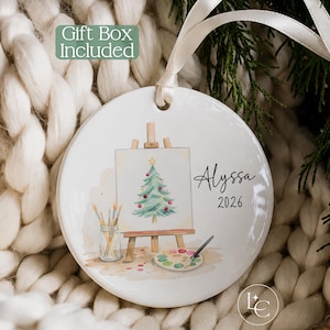 May include: A round white ceramic ornament featuring a watercolor illustration of a Christmas tree on an easel, a jar of paintbrushes, and a paint palette. The name Alyssa and the year 2026 are printed in black text. A white ribbon is attached.