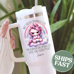 May include: White tumbler with a handle featuring a cartoon unicorn design. The unicorn has a pink mane and is sitting on a cloud under a rainbow. Text includes "NOT TODAY, SPRINKLE TITS" and "I'M RUNNIN' ON SPARKLES, SASS, AND ZERO F*CKS."