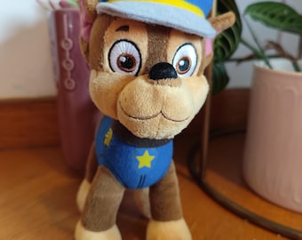 May include: A plush toy of a brown and tan dog wearing a blue and yellow police hat with a white paw print on it. The dog is wearing a blue uniform with a yellow star on the chest.