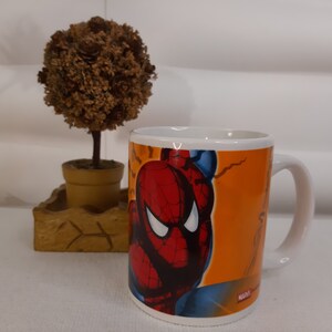 Op de afbeelding: Een witte keramische mok met een kleurrijk beeld van Spider-Man op een oranje achtergrond. De mok heeft een zwart handvat en het woord "MARVEL" staat op de bodem gedrukt.
