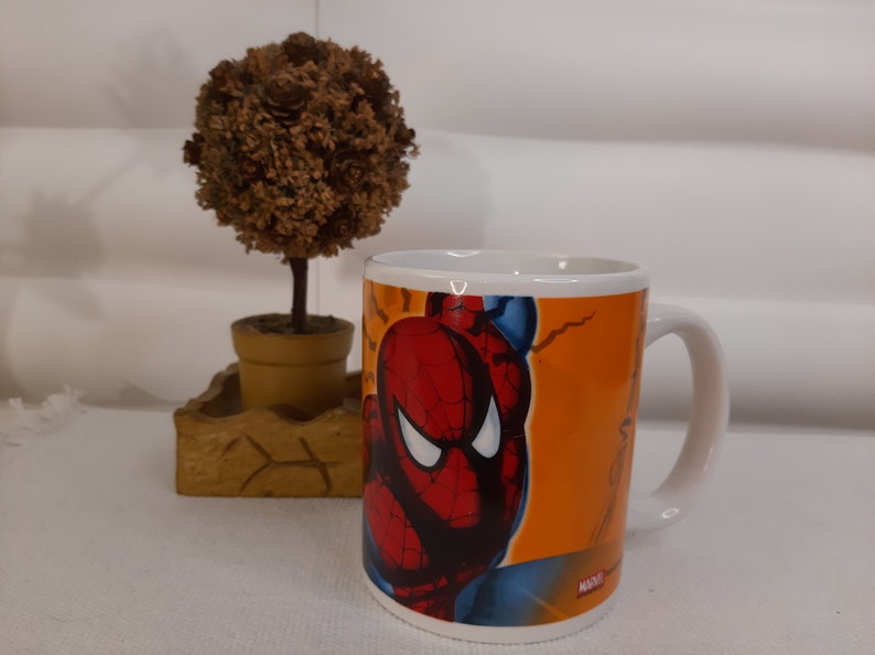 Op de afbeelding: Een witte keramische mok met een kleurrijk beeld van Spider-Man op een oranje achtergrond. De mok heeft een zwart handvat en het woord "MARVEL" staat op de bodem gedrukt.