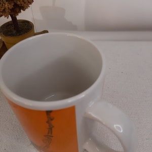 Op de afbeelding: Witte keramische mok met een oranje en zwart ontwerp. Het ontwerp toont een gestileerde afbeelding van een persoon met een gitaar.