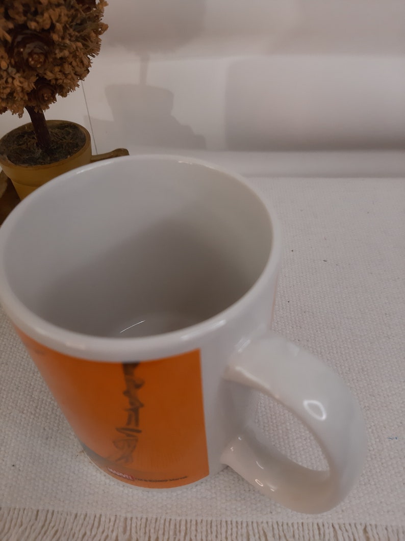 Op de afbeelding: Witte keramische mok met een oranje en zwart ontwerp. Het ontwerp toont een gestileerde afbeelding van een persoon met een gitaar.