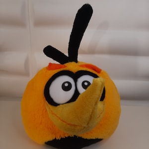 Puede incluir: Un peluche del personaje de Angry Birds, Chuck. El pájaro redondo y amarillo tiene un pico grande y amarillo, ojos grandes y blancos y detalles en negro. Tiene un mechón de plumas negras en la parte superior de la cabeza y una base negra.