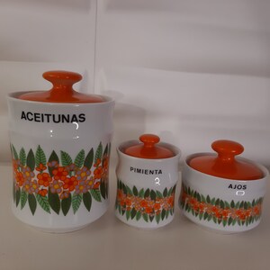Puede incluir: Tres botes de cerámica con tapas naranjas y diseños florales. Los botes están etiquetados con "Aceitunas", "Pimiento" y "Ajos".
