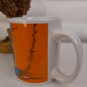 Op de afbeelding: Witte keramische mok met een oranje achtergrond en een zwart-witte afbeelding van een stripfiguur. De mok heeft een zwart handvat en de tekst "MARVEL TM & &copy;2009 Marvel." aan de onderkant.