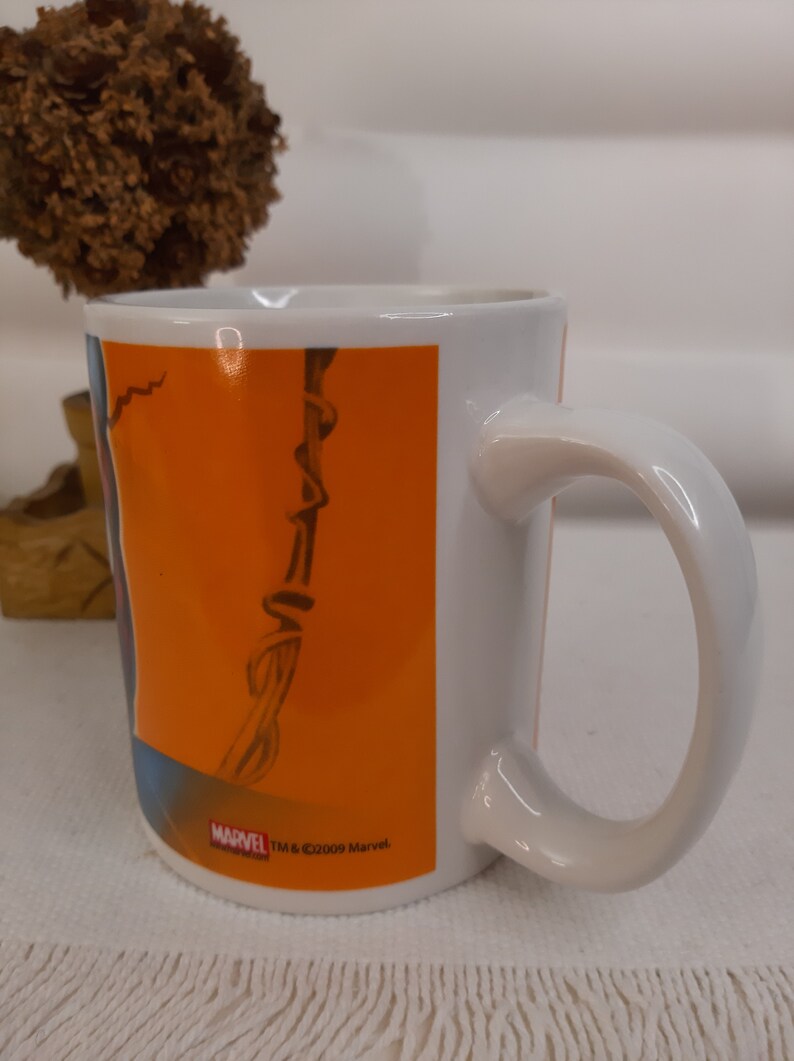 Op de afbeelding: Witte keramische mok met een oranje achtergrond en een zwart-witte afbeelding van een stripfiguur. De mok heeft een zwart handvat en de tekst "MARVEL TM & &copy;2009 Marvel." aan de onderkant.