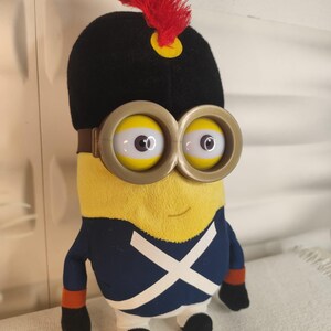 Könnte beinhalten: Ein Plüschtier-Minion in einer Uniform im napoleonischen Stil. Das Minion hat einen gelben Körper, schwarze Stiefel und eine blaue Jacke mit einem weißen Kreuz. Es trägt einen schwarzen Hut mit roter Feder und eine große, runde Brille.