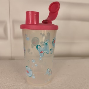 Puede incluir: Una botella de agua de plástico transparente con una tapa rosa y un pico abatible. La botella tiene un diseño de burbujas azules y peces rojos.