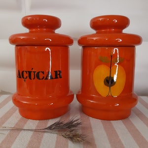Pode incluir: Dois potes de cerâmica laranja com tampas. Um pote tem a palavra "ACÚCAR" escrita em letras pretas. O outro pote tem uma imagem pintada de uma laranja cortada ao meio com folhas verdes.