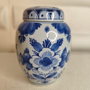 Może przedstawiać: Mały, biały ceramiczny słoik z pokrywką, ozdobiony niebieskimi wzorami kwiatowymi. Słoik ma zaokrąglony kształt i pasującą pokrywkę. Niebieskie wzory obejmują kwiaty, liście i inne elementy dekoracyjne. Słoik jest prawdopodobnie używany do przechowywania lub ekspozycji.