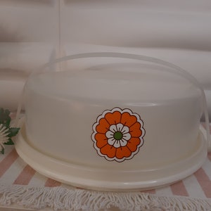 Pode incluir: Um porta-bolos branco com uma tampa transparente e um design floral laranja e branco. O porta-bolos tem uma alça na parte superior.