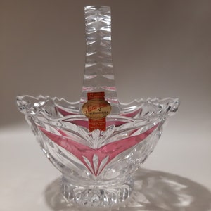 May include: A clear glass bowl with a pink rim and a cut glass handle. The bowl has a label that reads "Anna Hutte Bleikristall".