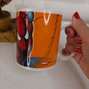 Op de afbeelding: Een witte keramische mok met een handvat. De mok heeft een kleurrijk beeld van Spiderman op een feloranje achtergrond. De tekst "MARVEL TM & &copy;2002 Marvel" staat gedrukt op de bodem van de mok.