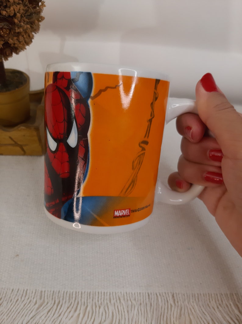 Op de afbeelding: Een witte keramische mok met een handvat. De mok heeft een kleurrijk beeld van Spiderman op een feloranje achtergrond. De tekst "MARVEL TM & &copy;2002 Marvel" staat gedrukt op de bodem van de mok.