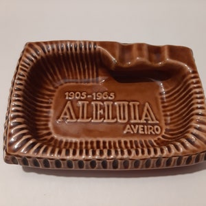 Op de afbeelding: Bruine asbak van keramiek met een verhoogde rand en een decoratief ontwerp. Het heeft de tekst "1905-1965 ALELUIA AVEIRO".