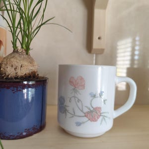 Puede incluir: Una taza de cerámica blanca con un diseño floral con flores rosas y azules y tallos verdes. La taza está sobre una estantería de madera junto a una maceta de cerámica azul con una pequeña planta verde.