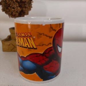 Op de afbeelding: Een witte keramische mok met een afbeelding van Spiderman. De mok heeft een oranje achtergrond met zwarte bliksemschichten en de woorden "Spider-Sense" en "Spider-Man" in zwarte tekst.