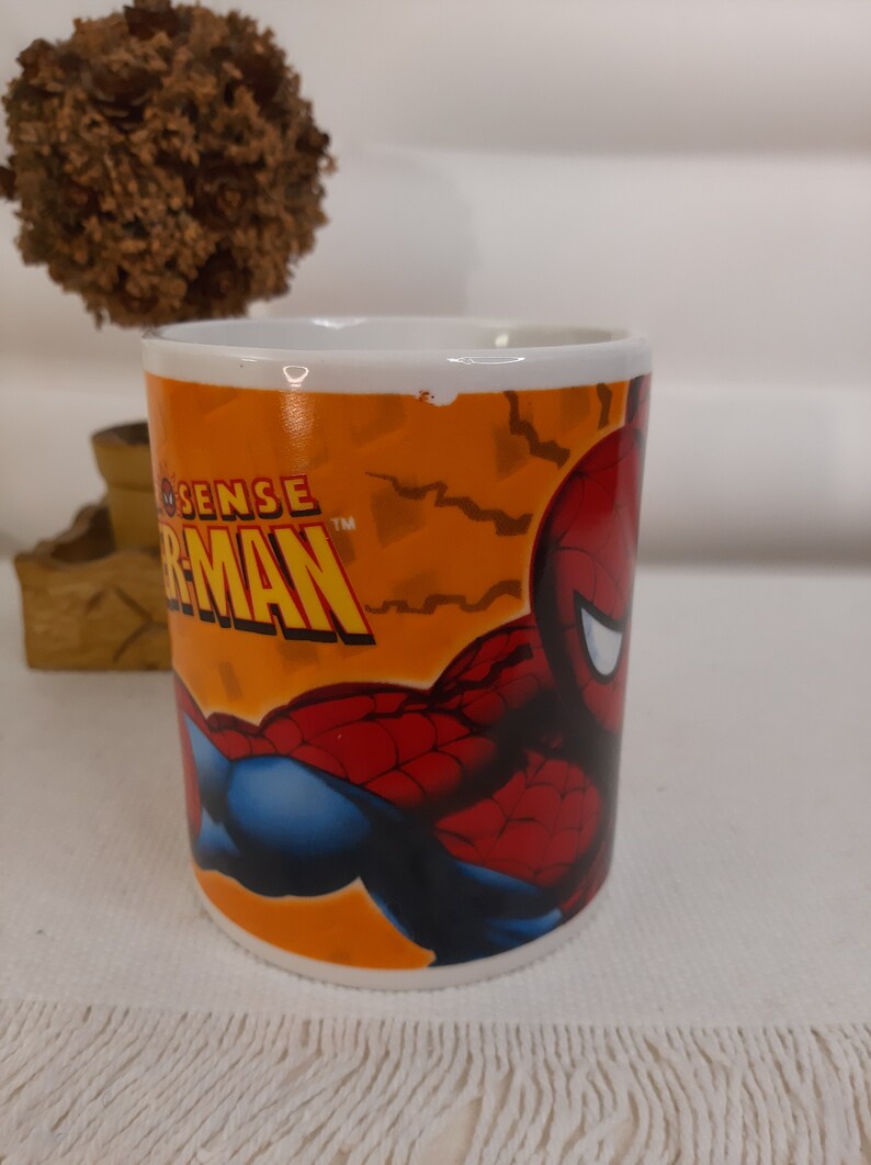 Op de afbeelding: Een witte keramische mok met een afbeelding van Spiderman. De mok heeft een oranje achtergrond met zwarte bliksemschichten en de woorden "Spider-Sense" en "Spider-Man" in zwarte tekst.