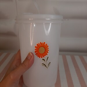 May include: White plastic tumbler with a lid and a straw. The tumbler has a floral design with an orange flower and green leaves.