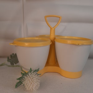 Puede incluir: Un especiero vintage blanco y amarillo con tres compartimentos, cada uno con una tapa amarilla. El especiero tiene un asa y una base amarillas. Una flor blanca y un tallo verde en primer plano.