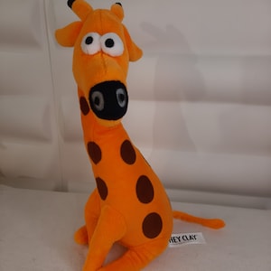 Puede incluir: Juguete de peluche de jirafa naranja con manchas marrones, nariz negra y ojos grandes con borde blanco. La jirafa está sentada con la cola extendida. La etiqueta dice "HEY CLAY".