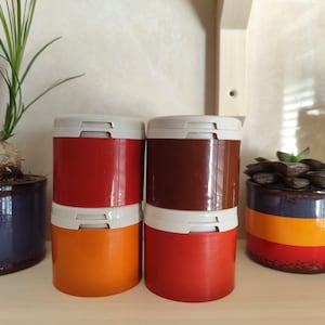 Pode incluir: Quatro recipientes cilíndricos em vermelho, laranja e castanho com tampas brancas estão empilhados. Duas plantas em vasos, uma com folhas verdes e outra com uma suculenta, estão em ambos os lados. Os vasos são azuis, vermelhos, amarelos e laranjas.