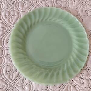 May include: A light green, vintage-style plate with a swirled rim design. The plate is round and has a smooth, glossy finish. The plate is made of glass and is a pale green color.