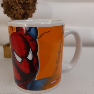 Op de afbeelding: Een witte keramische mok met een kleurrijk beeld van Spider-Man aan de voorkant. De mok heeft een zwarte omtrek rond het beeld en de woorden "MARVEL" in rode letters onderaan.