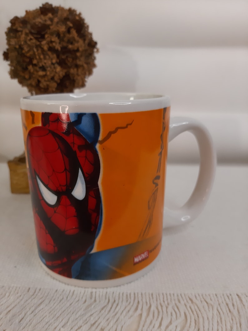 Op de afbeelding: Een witte keramische mok met een kleurrijk beeld van Spider-Man aan de voorkant. De mok heeft een zwarte omtrek rond het beeld en de woorden "MARVEL" in rode letters onderaan.