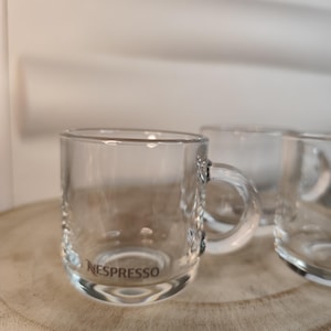 Peut inclure: Trois tasses à espresso en verre transparent avec une poignée. La tasse au premier plan porte l'inscription "NESPRESSO" au fond.